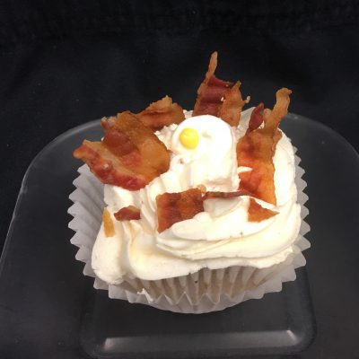 A bacon caramel cupcake topped with maple buttercream, bacon pieces, and a candy egg. Bacon and Eggs