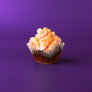A red velvet cupcake with a peanut butter ganache filling, topped with a white chocolate buttercream icing, and finished with white morsels and a peanut butter drizzle. Peanut Butter Blush cupcake