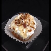 A cinnamon roll cupcake with bacon baked in, topped with vanilla buttercream, and finished with a cinnamon roll glaze drizzle, cinnamon sugar sprinkles, and bacon pieces. Bacon Cinnamon Roll cupcake