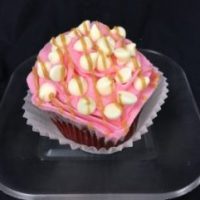 A red velvet cupcake with a peanut butter ganache filling, topped with a white chocolate buttercream icing, and finished with white morsels and a peanut butter drizzle. Peanut Butter Blush cupcake