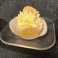 A banana cupcake with white ganache filling, iced with banana buttercream and topped with a white ganache drizzle, white morsels, and a banana chip. White Chocolate Banana cupcake